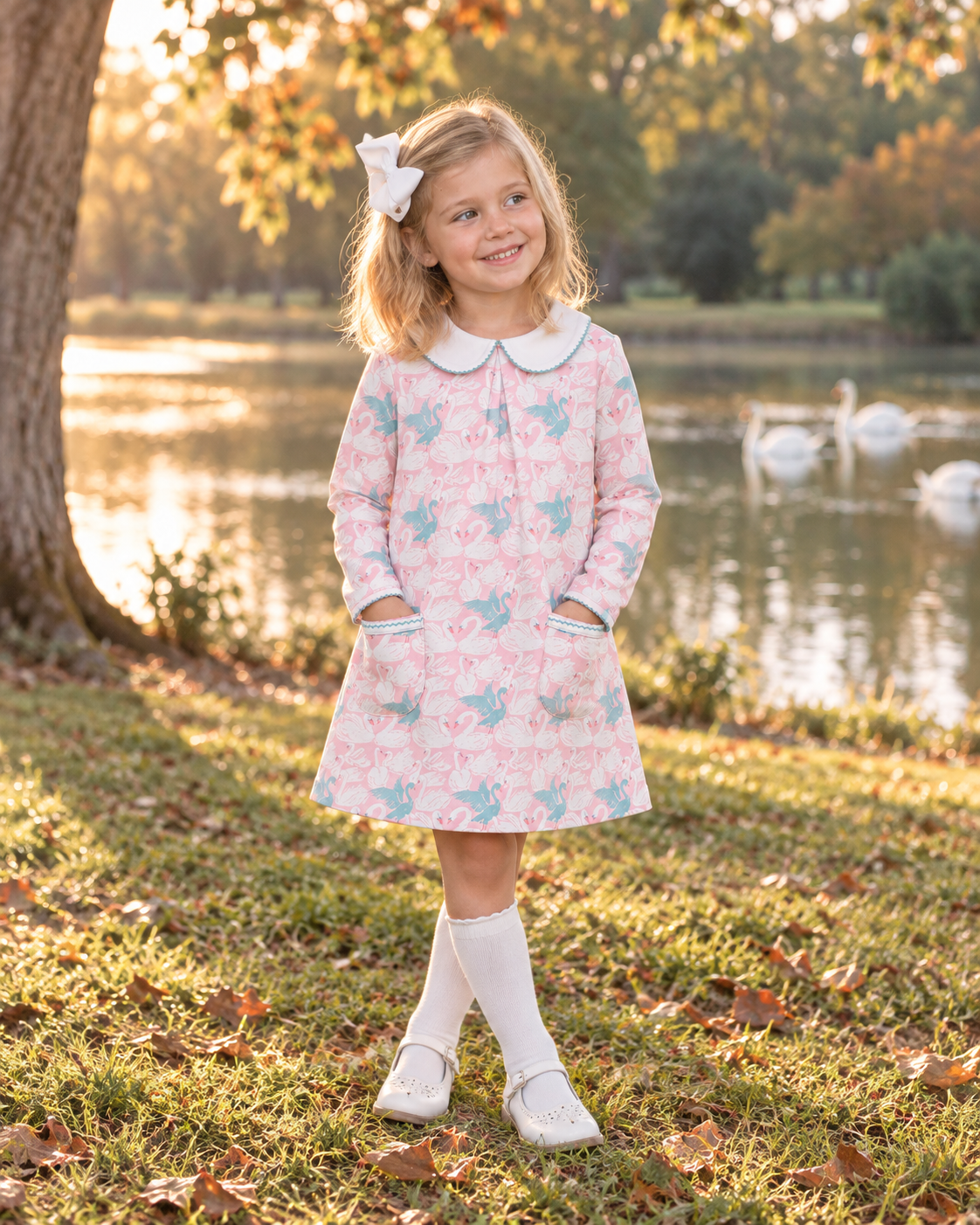 A young girl stands on a grassy park path by a lake, wearing a light pink long-sleeve dress with a white Peter Pan collar and a blue and pink pattern of small birds and hearts. She is wearing white knee-high socks and white shoes, with a white bow in her hair.