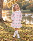 A young girl stands on a grassy park path by a lake, wearing a light pink long-sleeve dress with a white Peter Pan collar and a blue and pink pattern of small birds and hearts. She is wearing white knee-high socks and white shoes, with a white bow in her hair.