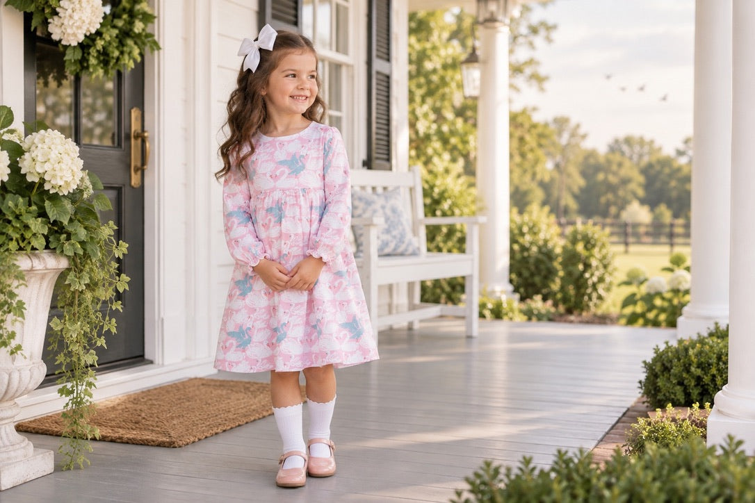A young girl stands on a front porch wearing a pink long-sleeve dress with a light blue floral pattern, white socks, and pink shoes. The background shows a white house, columns, and greenery.