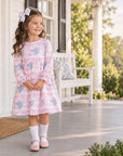 A young girl stands on a front porch wearing a pink long-sleeve dress with a light blue floral pattern, white socks, and pink shoes. The background shows a white house, columns, and greenery.