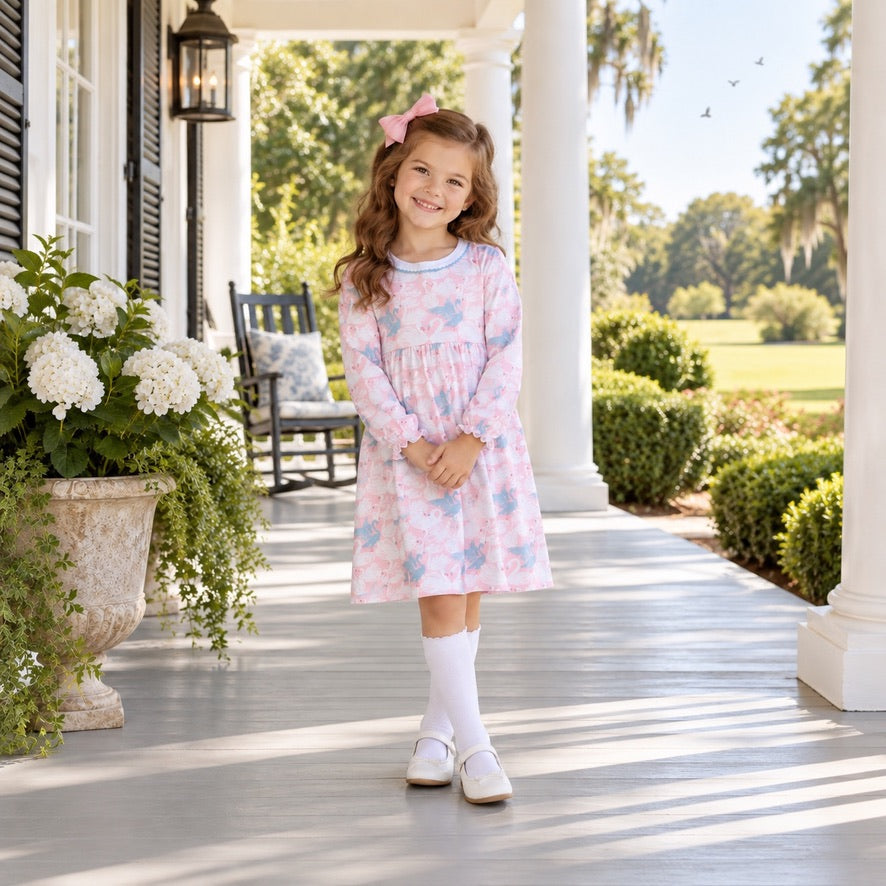 A young girl stands on a sunlit porch wearing a light pink long-sleeve dress with a blue and white floral print, white knee-high socks, and white shoes. The background shows white columns, a rocking chair, large planters with white flowers, and a green lawn.