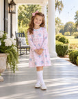 A young girl stands on a sunlit porch wearing a light pink long-sleeve dress with a blue and white floral print, white knee-high socks, and white shoes. The background shows white columns, a rocking chair, large planters with white flowers, and a green lawn.