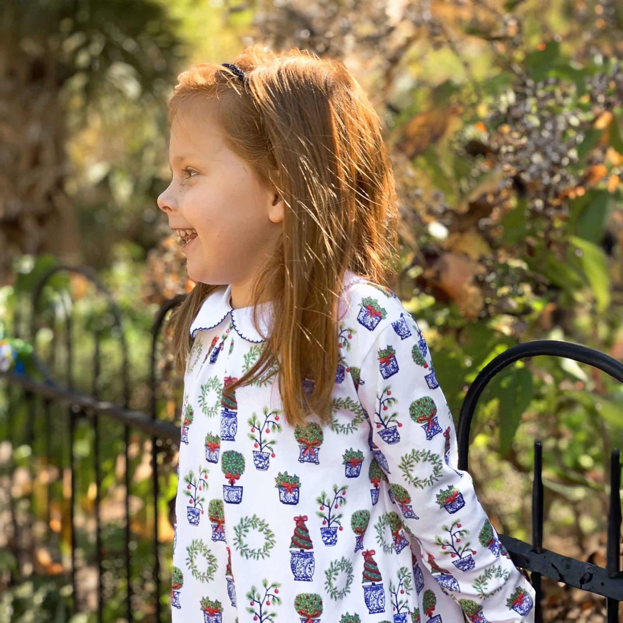 redhead girl leaning on fence wearing white, blue, red and green dress with christmas topiaries 