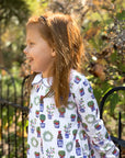 redhead girl leaning on fence wearing white, blue, red and green dress with christmas topiaries 