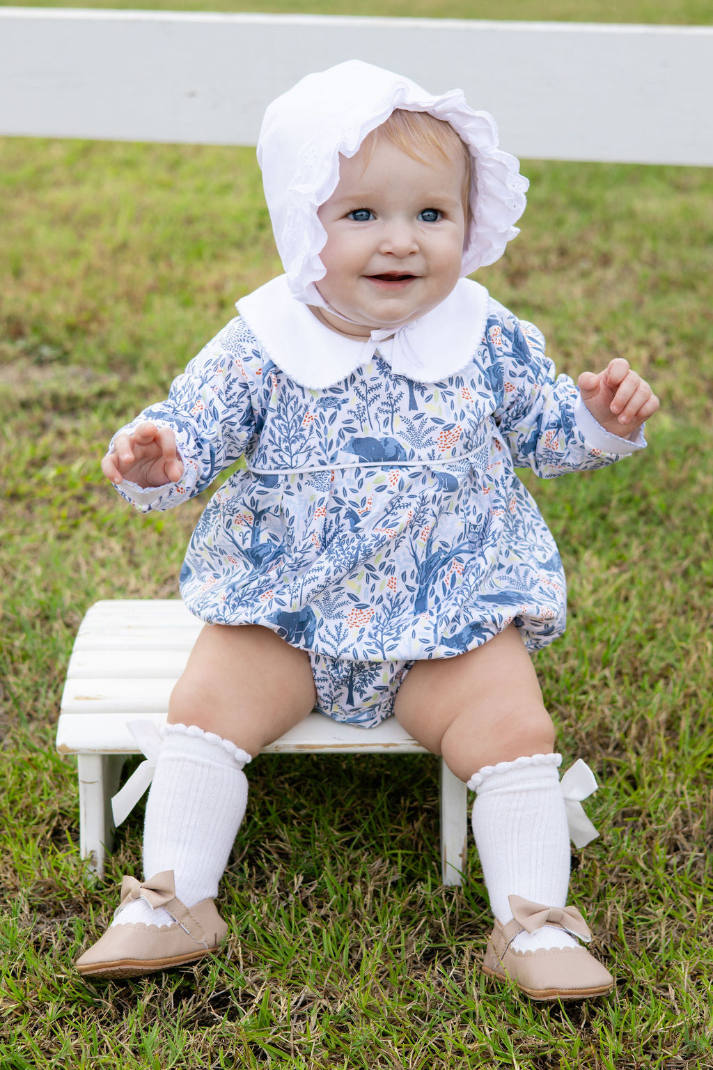 Baby sitting on a small stool outdoors wearing a floral romper and bonnet.