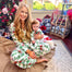 A woman sitting on a rug wearing a Christmas-themed pajama set with a child in her lap, in a room with a Christmas tree and holiday decorations in the background.