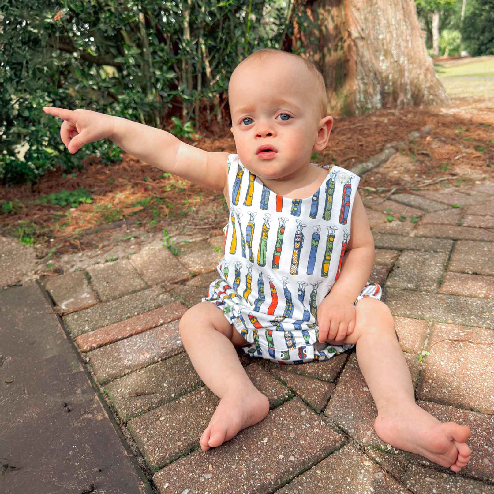 Baby sitting on a paved walkway pointing to the side, wearing a colorful golf club bubble.