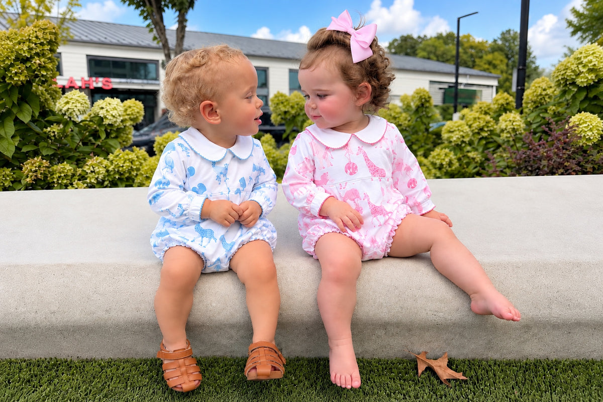 Two toddlers sitting on a concrete bench outdoors, wearing matching printed romper outfits. The child on the left wears a light blue and white patterned romper with a white collar and brown sandals.