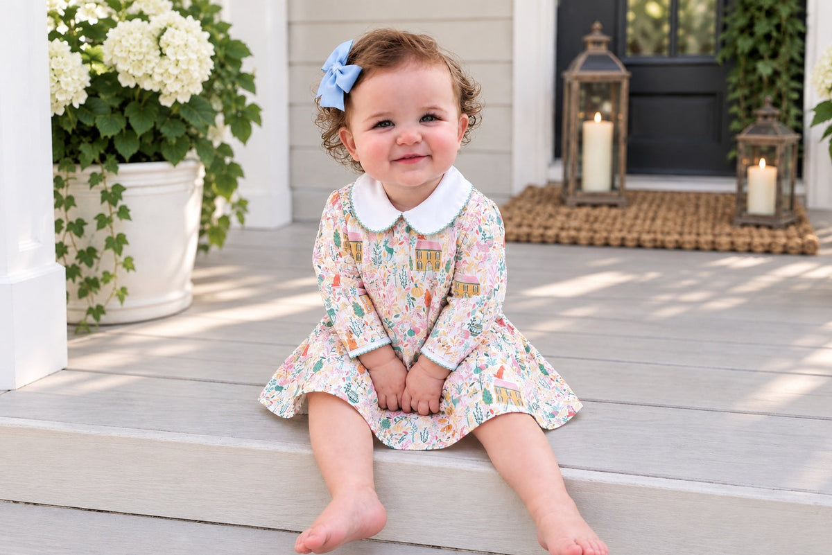 A toddler girl sits barefoot on the steps of a front porch wearing a long-sleeve floral dress with a white Peter Pan collar and a blue bow in her hair.