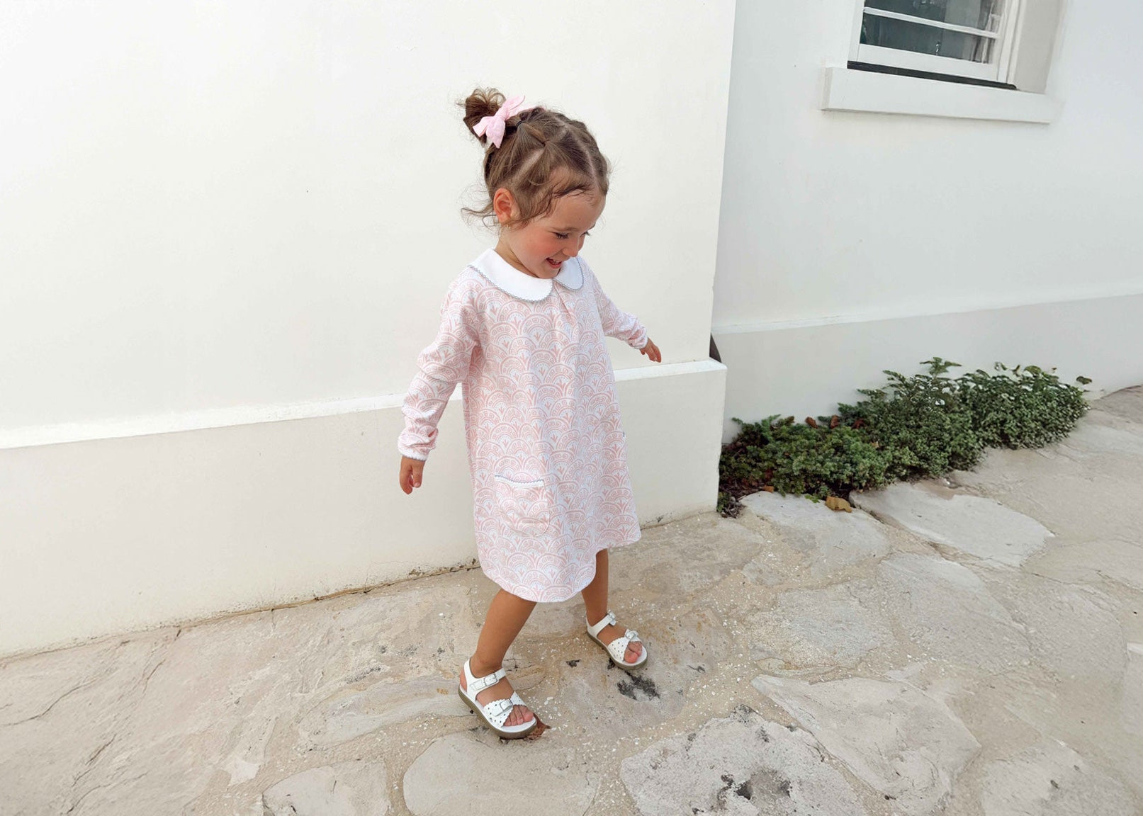 Young girl in a white and pink scallop pattern dress standing on a stone path next to a white wall.