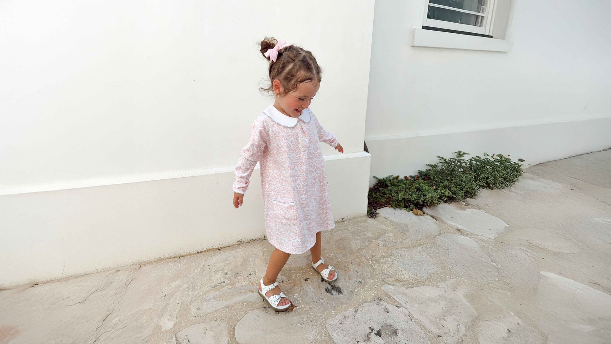 Young girl in a white and pink scallop pattern dress standing on a stone path next to a white wall.