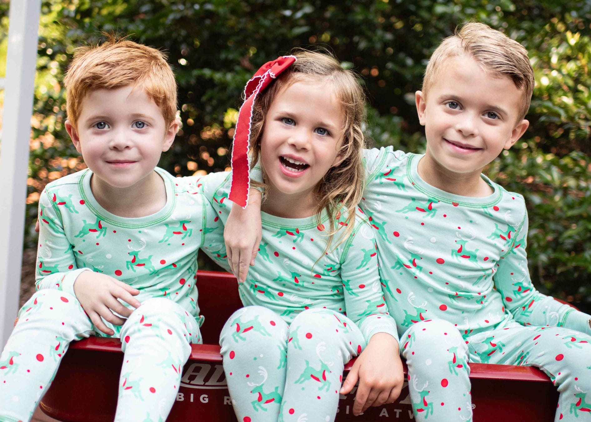 Three children in matching pajamas sitting in a red wagon outdoors.