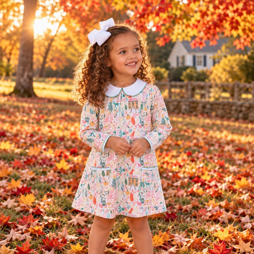 A young girl with curly brown hair wearing a long-sleeve floral dress with a white Peter Pan collar and a white bow in her hair, standing in a yard covered with fallen autumn leaves.