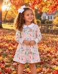 A young girl with curly brown hair wearing a long-sleeve floral dress with a white Peter Pan collar and a white bow in her hair, standing in a yard covered with fallen autumn leaves.
