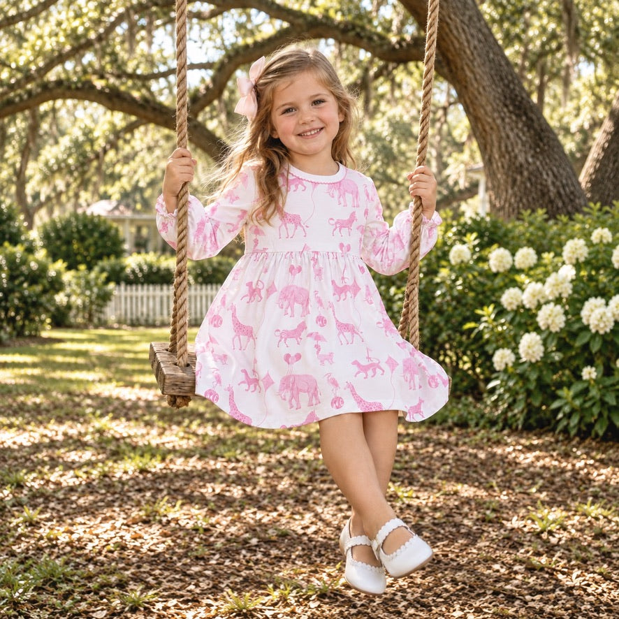 A young girl with a pink bow sits on a rope swing in a sunny backyard garden, wearing a white long-sleeve dress with a pink giraffe and heart pattern.