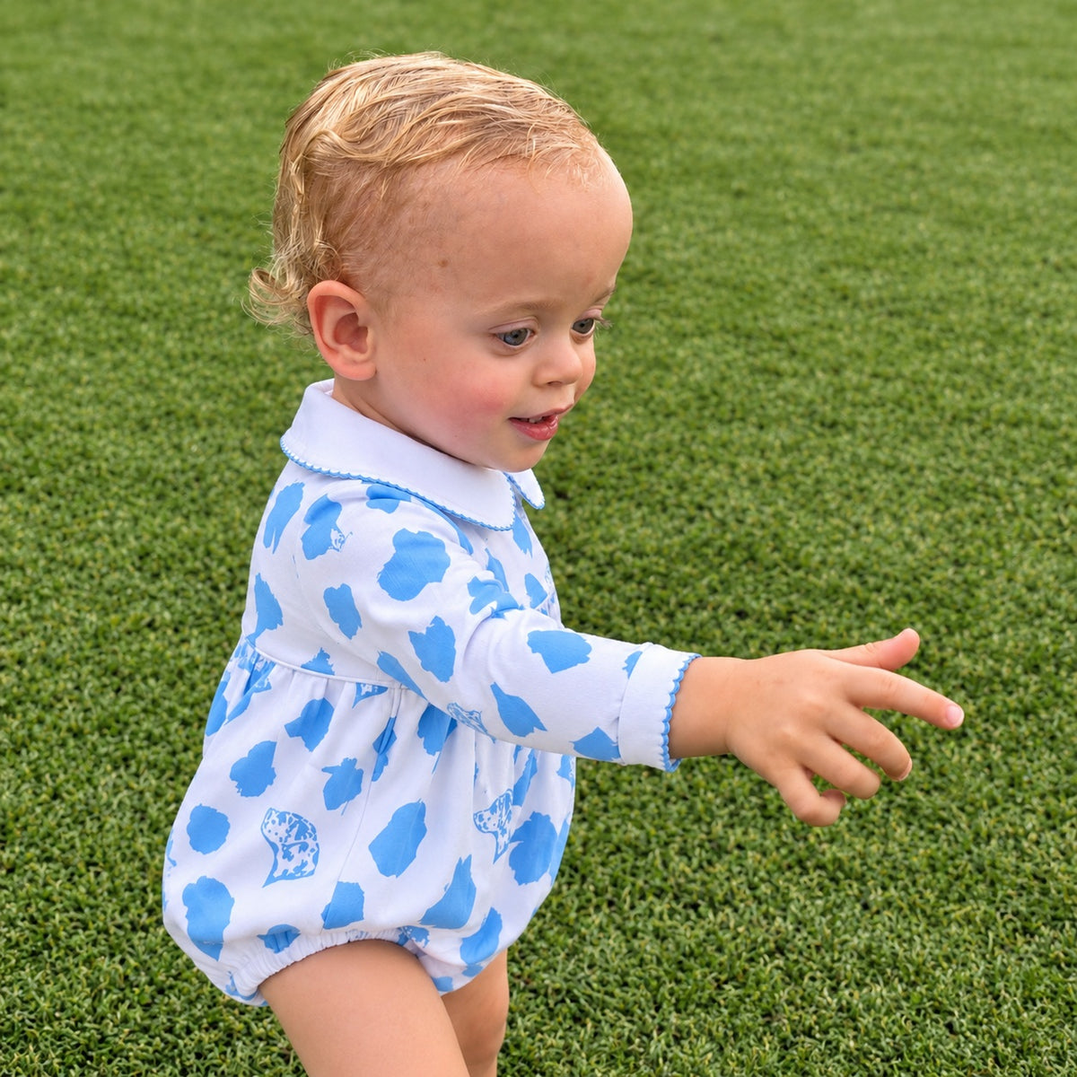 A toddler boy wearing a white long-sleeve romper with blue cloud patterns and a white collar, standing on green grass and pointing to the side.