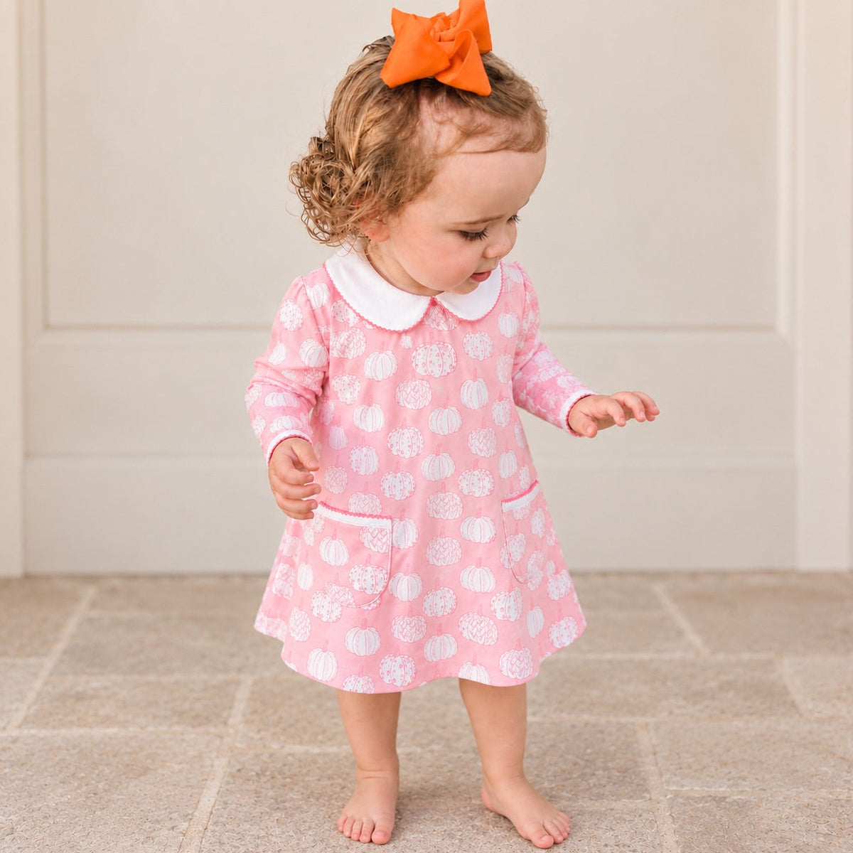 A toddler girl wearing a pink long-sleeve dress with a white pumpkin pattern, a white Peter Pan collar, and front pockets, standing barefoot on a tiled floor in front of a white paneled wall.