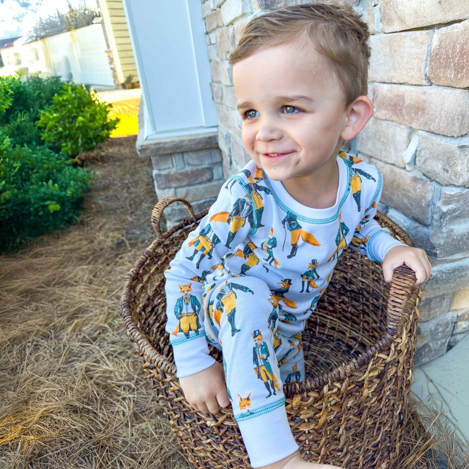 Child wearing pajamas with a fox pattern, sitting in a wicker basket outdoors.