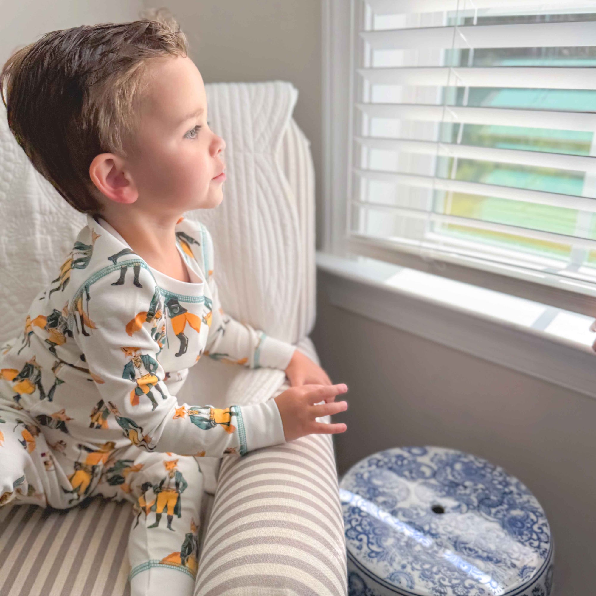 Child in pajamas sitting on a chair by a window with blinds.