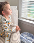Child in pajamas sitting on a chair by a window with blinds.